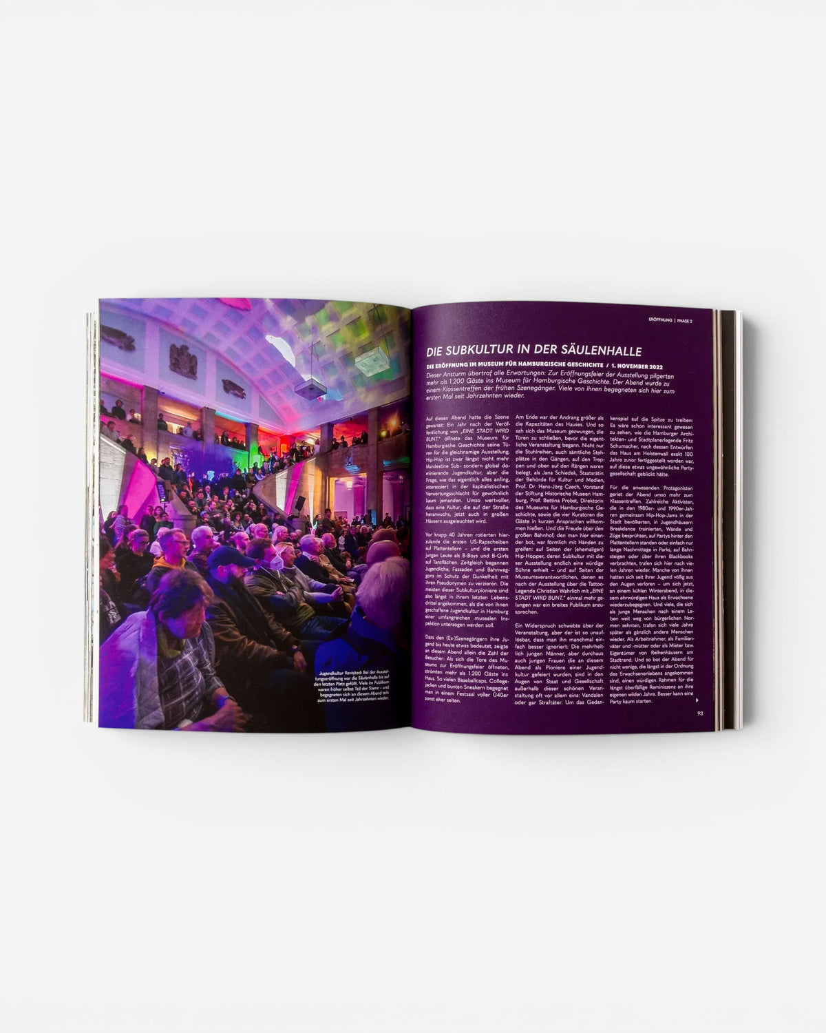 Open pages of Astrofat book EINE STADT WIRD BUNT. showing a crowded event in a hall with purple lighting and text.